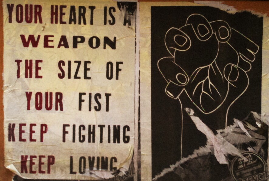 wheatpasted street art. on the left, text in all caps stating "your heart is a weapon the size of your fist keep loving keep fighting". on the right a white drawing of a hand holding a heart on a black background.